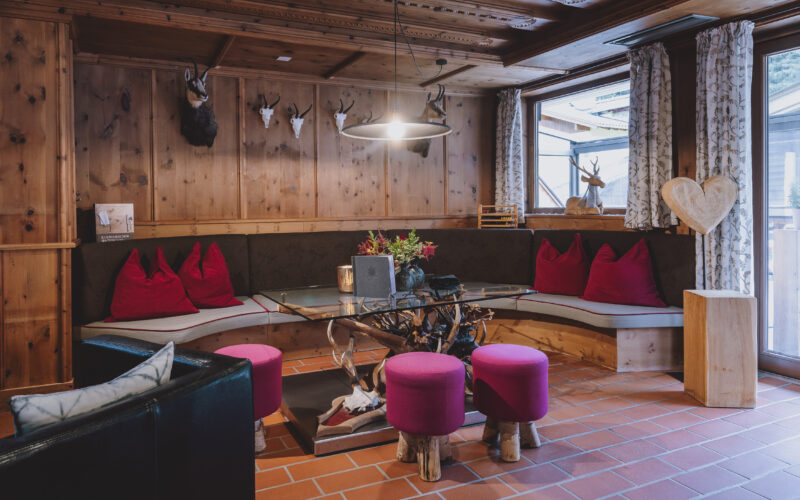 Gemütliche Sitzecke aus Holz mit einer geschwungenen Bank, roten Kissen, runden Hockern, einem gläsernen Couchtisch, Geweihdekor und natürlichem Licht durch ein großes Fenster.