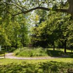Ein sonnenbeschienener Park mit einem Kiesweg, der sich um einen grasbewachsenen, kreisförmigen Bereich windet, umgeben von hohen Bäumen und einem Schild auf der linken Seite.