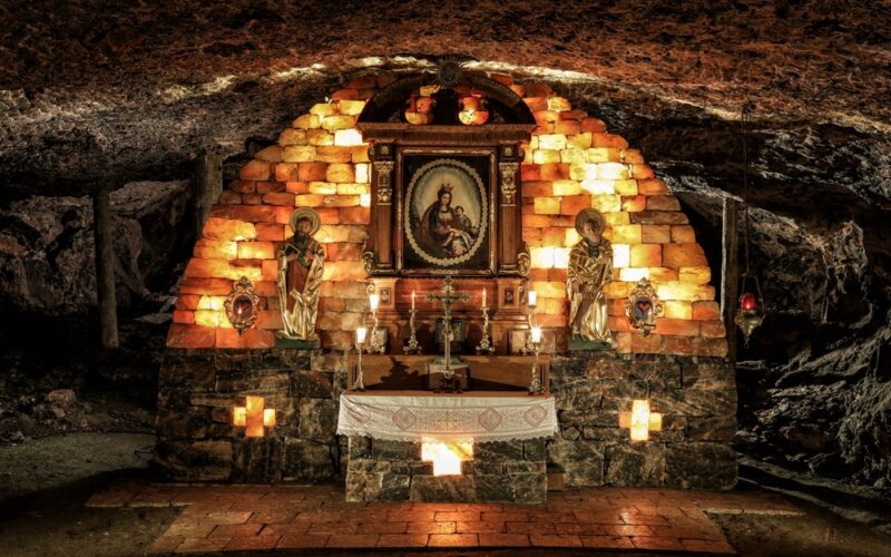 Eine unterirdische Kapelle mit einem Steinaltar, religiösen Statuen, brennenden Kerzen und einem gerahmten Gemälde in der Mitte, das an einer gewölbten Ziegelwand hängt.