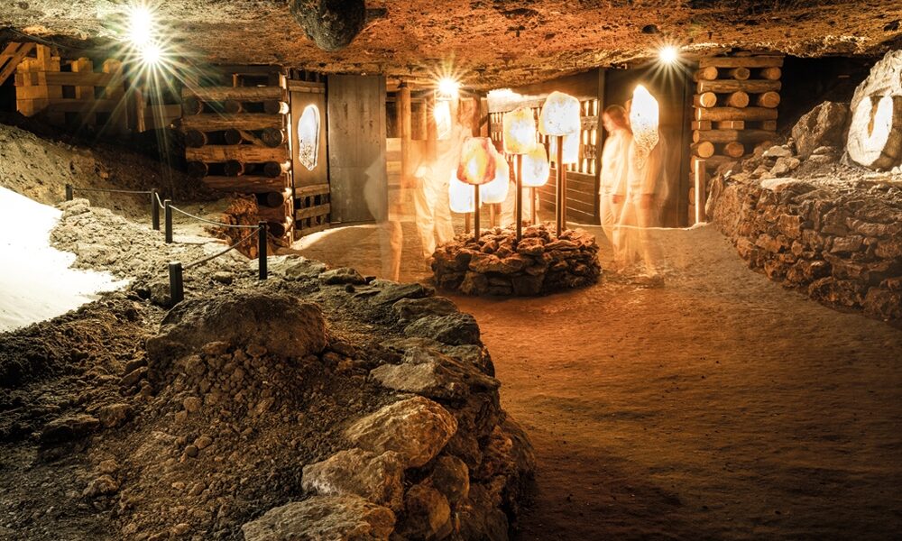 Schummerig beleuchtete unterirdische Höhle mit felsigen Wänden, freiliegenden Holzstützen und beleuchteten Figuren, die in der Mitte neben großen Salzkristallen stehen.