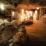 Schummerig beleuchtete unterirdische Höhle mit felsigen Wänden, freiliegenden Holzstützen und beleuchteten Figuren, die in der Mitte neben großen Salzkristallen stehen.