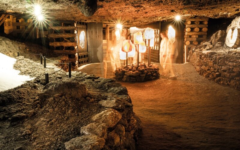Schummerig beleuchtete unterirdische Höhle mit felsigen Wänden, freiliegenden Holzstützen und beleuchteten Figuren, die in der Mitte neben großen Salzkristallen stehen.