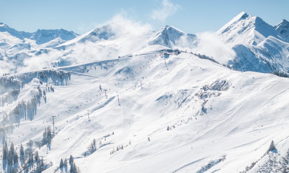 Schneebedeckte Berglandschaft mit Skipisten, Sesselliften, vereinzelten Bäumen und entfernten Gipfeln unter einem klaren blauen Himmel.