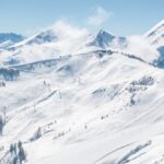 Schneebedeckte Berglandschaft mit Skipisten, Sesselliften, vereinzelten Bäumen und entfernten Gipfeln unter einem klaren blauen Himmel.