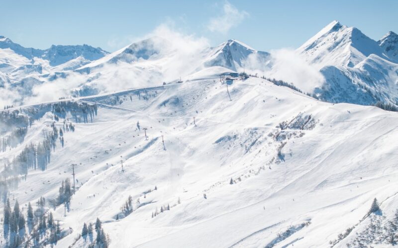 Schneebedeckte Berglandschaft mit Skipisten, Sesselliften, vereinzelten Bäumen und entfernten Gipfeln unter einem klaren blauen Himmel.