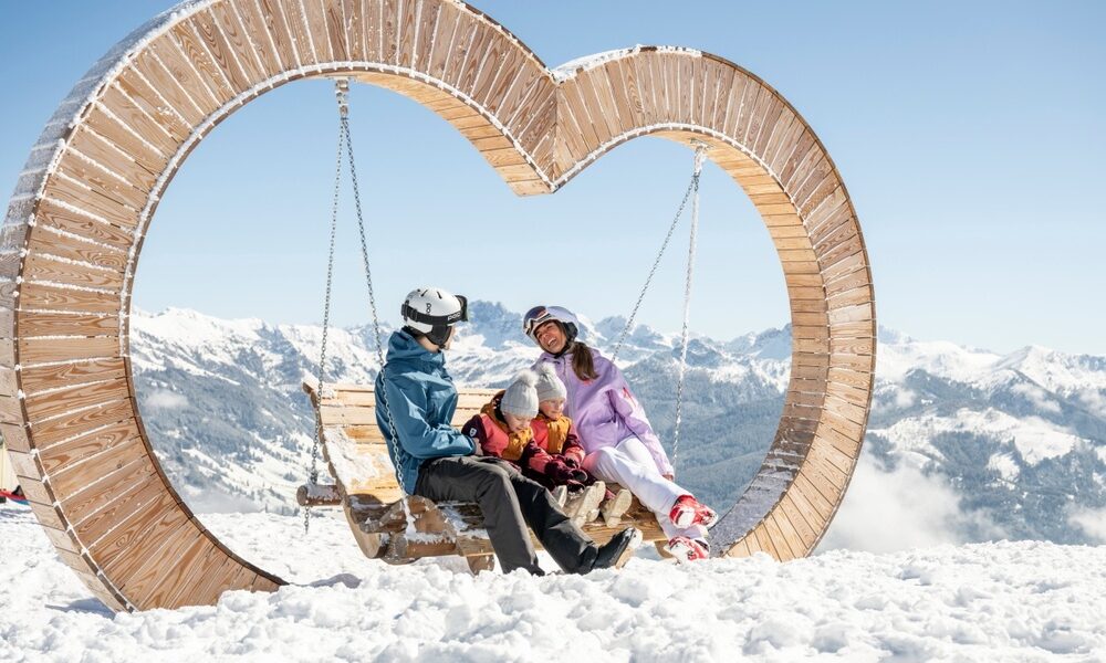 Eine vierköpfige Familie sitzt auf einer hölzernen Schaukel in Form eines Herzens, umgeben von Schnee und Bergen unter einem strahlend blauen Himmel.