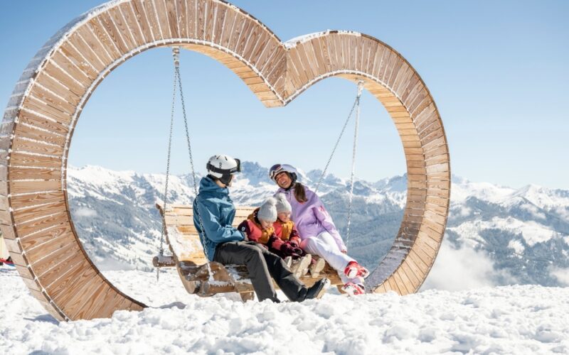 Eine vierköpfige Familie sitzt auf einer hölzernen Schaukel in Form eines Herzens, umgeben von Schnee und Bergen unter einem strahlend blauen Himmel.