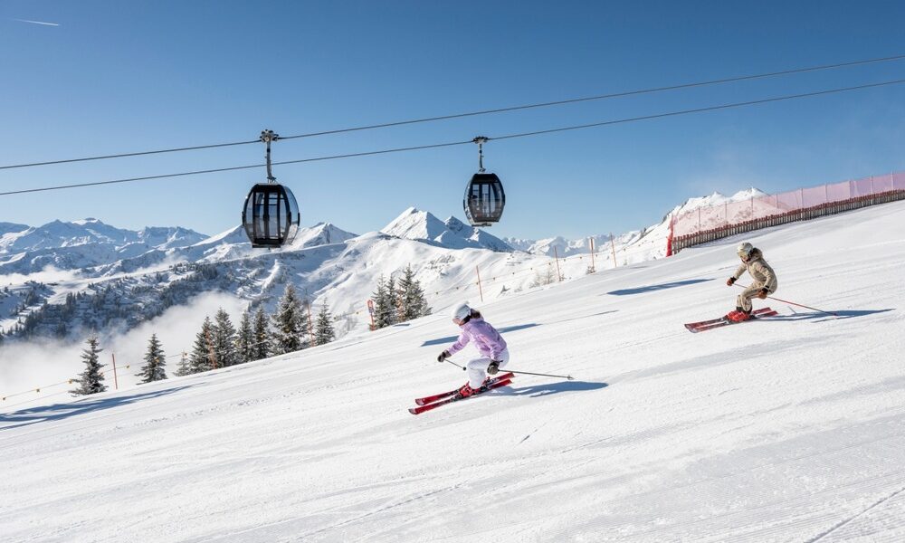 Zwei Skifahrer gleiten unter den Gondeln eines Skilifts einen verschneiten Hang hinunter, mit Bergen und Kiefern im Hintergrund an einem klaren Tag.