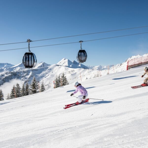 Zwei Skifahrer gleiten unter den Gondeln eines Skilifts einen verschneiten Hang hinunter, mit Bergen und Kiefern im Hintergrund an einem klaren Tag.