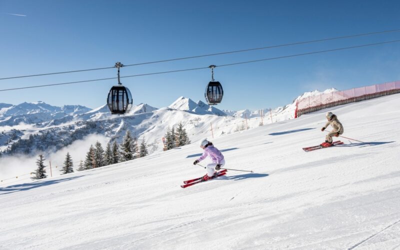 Zwei Skifahrer gleiten unter den Gondeln eines Skilifts einen verschneiten Hang hinunter, mit Bergen und Kiefern im Hintergrund an einem klaren Tag.