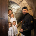 Ein Mann in schwarzer historischer Uniform steht an einem Tunneleingang und gestikuliert dorthin, während eine lächelnde Familie mit zwei Kindern in weißen Mänteln an der Tür steht.