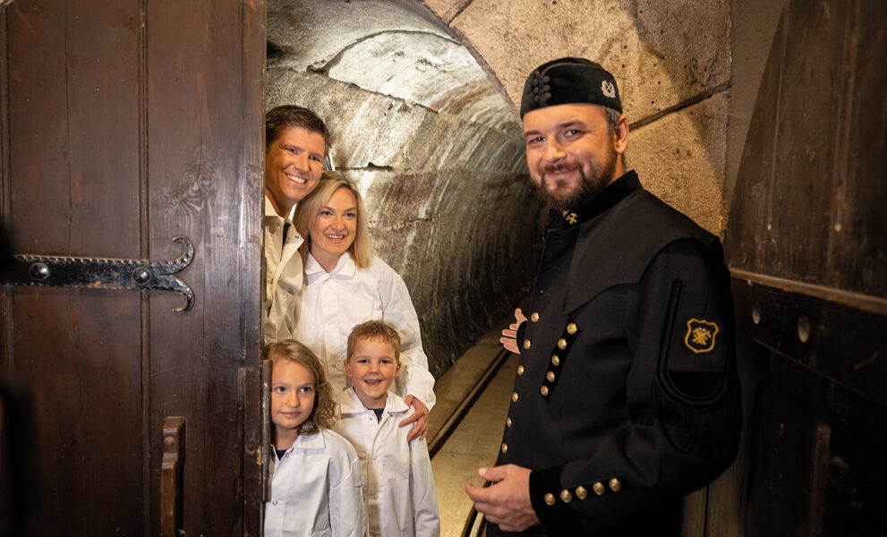 Ein Mann in schwarzer historischer Uniform steht an einem Tunneleingang und gestikuliert dorthin, während eine lächelnde Familie mit zwei Kindern in weißen Mänteln an der Tür steht.