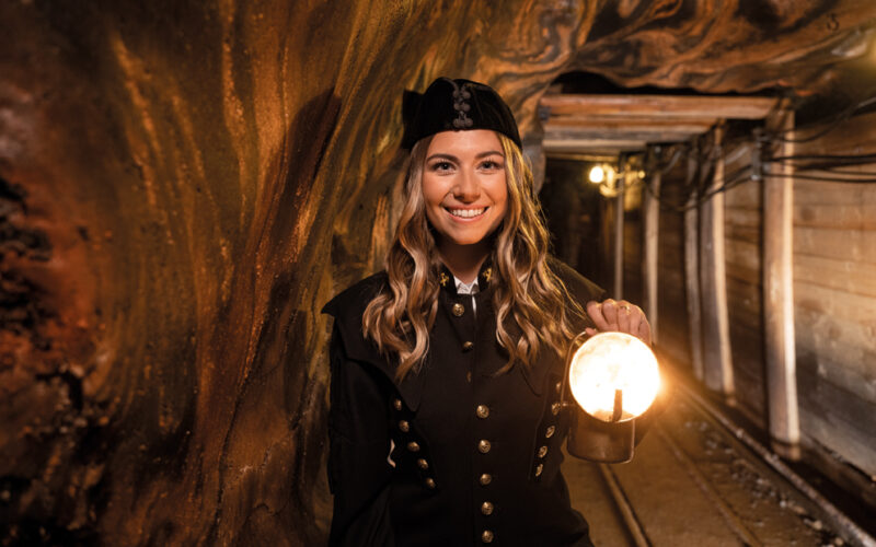 Eine Frau in schwarzer Uniform hält eine brennende Laterne, während sie in einem unterirdischen Stollen mit Holzstützen steht.