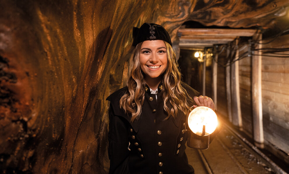 Eine Frau in schwarzer Uniform hält eine brennende Laterne, während sie in einem unterirdischen Stollen mit Holzstützen steht.