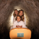 Drei weiß gekleidete Personen sitzen in einer Reihe in einer engen Höhle oder einem Tunnel, vor ihnen ein Holzsitz mit der Aufschrift "6".