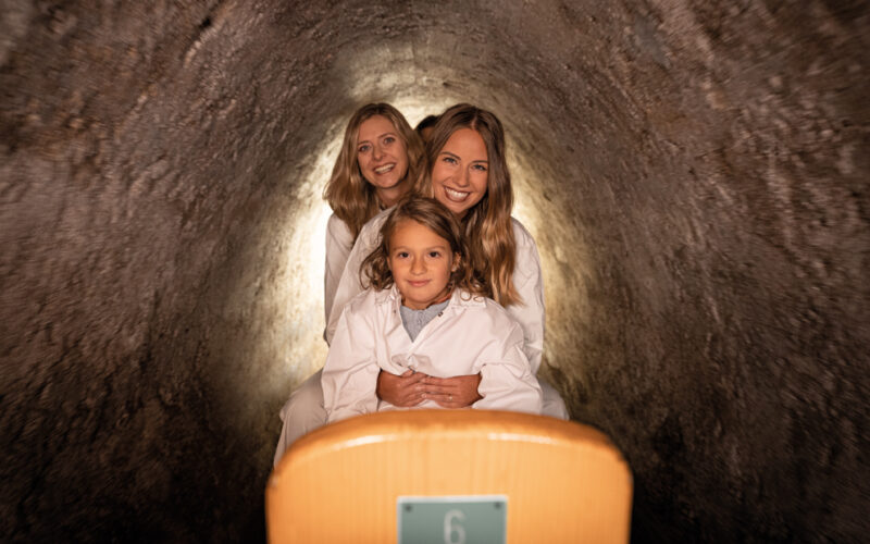 Drei weiß gekleidete Personen sitzen in einer Reihe in einer engen Höhle oder einem Tunnel, vor ihnen ein Holzsitz mit der Aufschrift "6".
