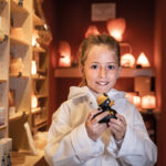 Ein junges Mädchen in einem weißen Mantel lächelt und hält eine kleine schwarze Figur in einem warm beleuchteten Geschäft mit Regalen mit verschiedenen Produkten im Hintergrund.