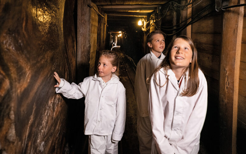 Drei Kinder in weißen Schutzanzügen gehen durch einen schwach beleuchteten unterirdischen Tunnel mit Holzwänden und schauen sich neugierig um.