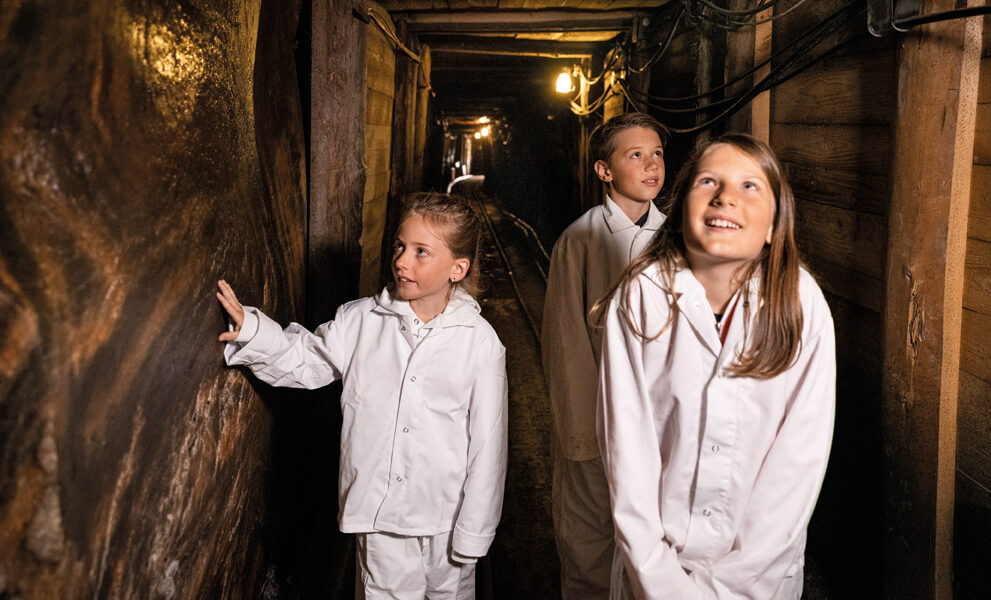 Drei Kinder in weißen Schutzanzügen gehen durch einen schwach beleuchteten unterirdischen Tunnel mit Holzwänden und schauen sich neugierig um.