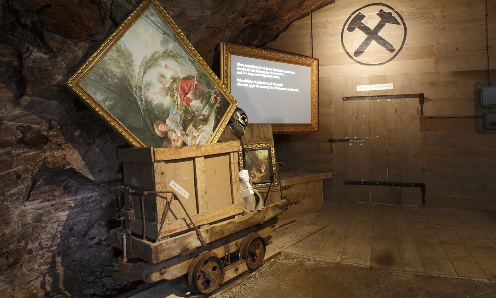 Ein hölzerner Bergbauwagen mit gerahmten Gemälden steht in einem schwach beleuchteten Raum mit Holzwänden, einem Bergbausymbol und einer beleuchteten Informationstafel im Hintergrund.