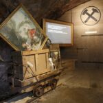 Ein hölzerner Bergbauwagen mit gerahmten Gemälden steht in einem schwach beleuchteten Raum mit Holzwänden, einem Bergbausymbol und einer beleuchteten Informationstafel im Hintergrund.