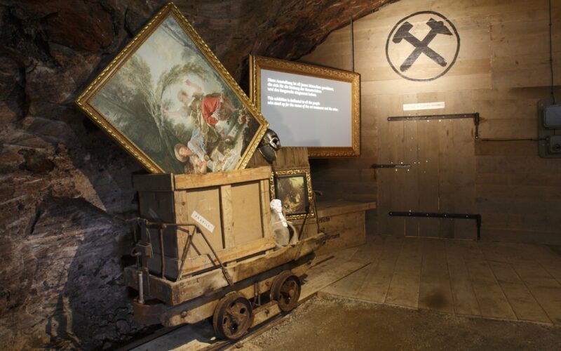 Ein hölzerner Bergbauwagen mit gerahmten Gemälden steht in einem schwach beleuchteten Raum mit Holzwänden, einem Bergbausymbol und einer beleuchteten Informationstafel im Hintergrund.