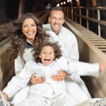 Ein Kind und zwei Erwachsene in weißen Kleidern rutschen gemeinsam eine Holzrutsche hinunter, lächeln und scheinen begeistert zu sein.