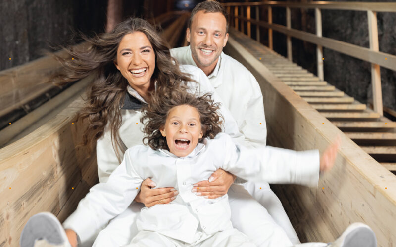 Ein Kind und zwei Erwachsene in weißen Kleidern rutschen gemeinsam eine Holzrutsche hinunter, lächeln und scheinen begeistert zu sein.