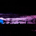 Eine große blaue Kugel und verschiedene Objekte werden von lila und rosa Lichtern in einer felsigen, höhlenartigen unterirdischen Umgebung beleuchtet.