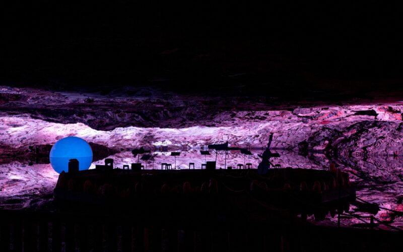 Eine große blaue Kugel und verschiedene Objekte werden von lila und rosa Lichtern in einer felsigen, höhlenartigen unterirdischen Umgebung beleuchtet.