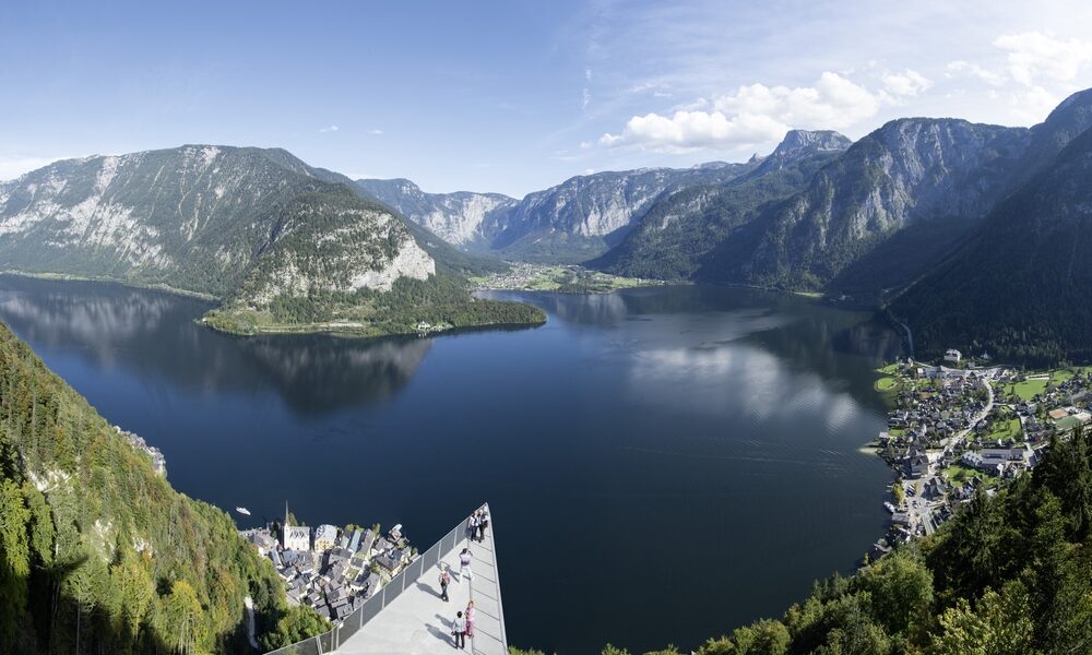 Panoramablick auf einen von Bergen umgebenen See mit einer Aussichtsplattform im Vordergrund und einem kleinen Dorf am Seeufer.