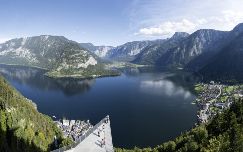 Panoramablick auf einen von Bergen umgebenen See mit einer Aussichtsplattform im Vordergrund und einem kleinen Dorf am Seeufer.