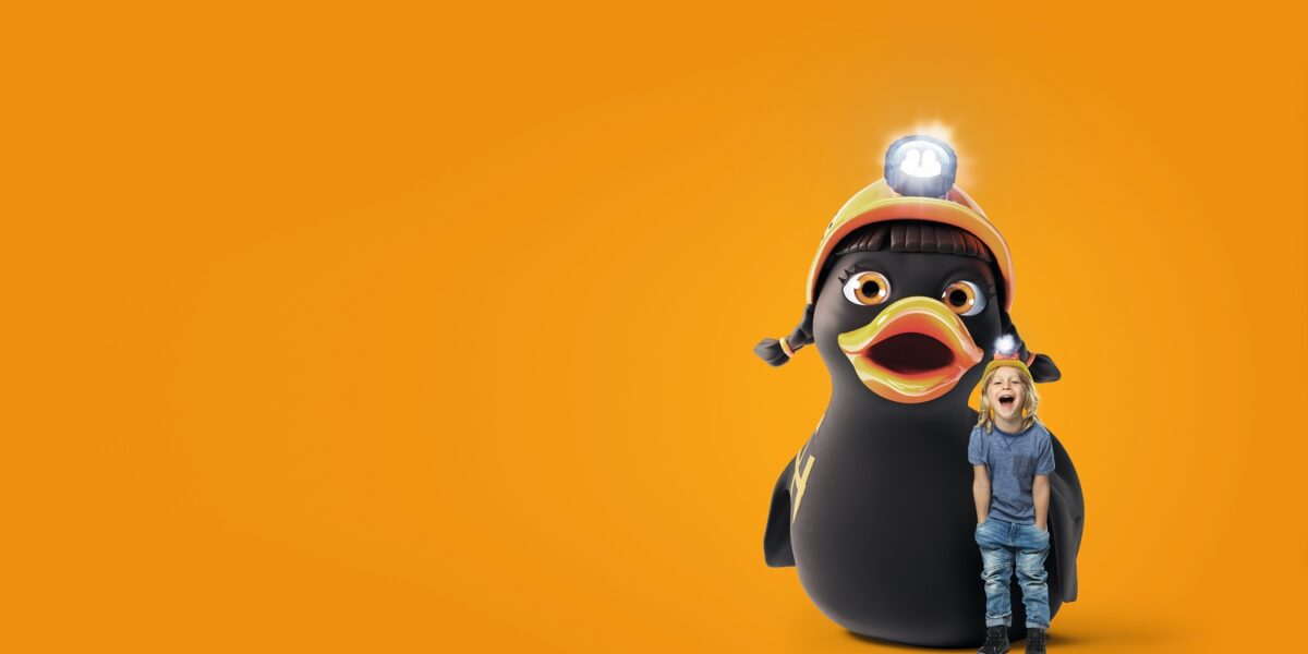 Ein kleines Kind steht neben einem großen Cartoon-Pinguin, der einen Helm mit einer Stirnlampe trägt, vor einem leuchtend orangefarbenen Hintergrund.