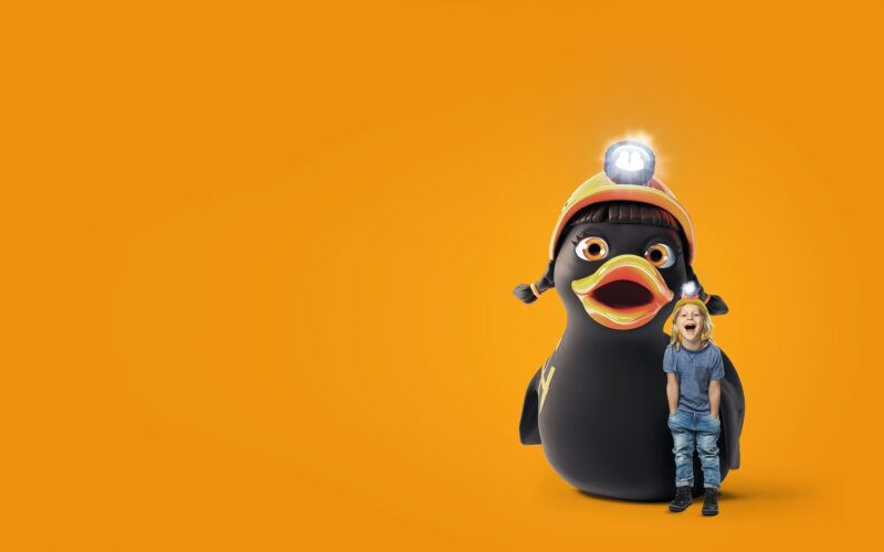 Ein kleines Kind steht neben einem großen Cartoon-Pinguin, der einen Helm mit einer Stirnlampe trägt, vor einem leuchtend orangefarbenen Hintergrund.