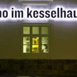Weiße Gebäudefassade bei Nacht mit einem beleuchteten Schild mit der Aufschrift "kino im kesselhaus" über einem Fenster, das einen Blick auf einen Raum mit verschiedenen Gegenständen im Inneren zeigt.