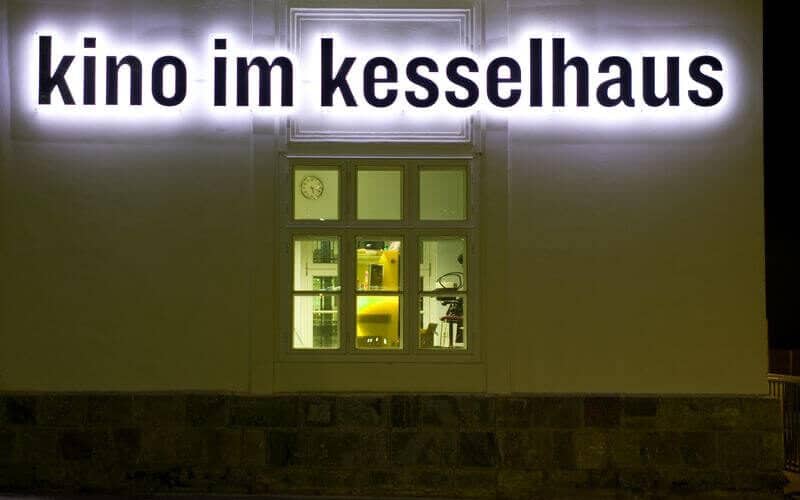 Weiße Gebäudefassade bei Nacht mit einem beleuchteten Schild mit der Aufschrift "kino im kesselhaus" über einem Fenster, das einen Blick auf einen Raum mit verschiedenen Gegenständen im Inneren zeigt.