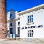 Weißes Gebäude mit der Aufschrift "kino im kesselhaus" an der Wand, daneben ein hoher Backsteinschornstein, unter blauem Himmel.