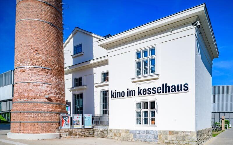 Weißes Gebäude mit der Aufschrift "kino im kesselhaus" an der Wand, daneben ein hoher Backsteinschornstein, unter blauem Himmel.
