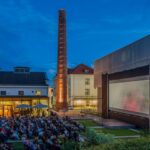 Eine große Gruppe von Menschen sieht sich in der Abenddämmerung einen Film auf einer Freiluftleinwand in einem Innenhof an, der von modernen Gebäuden und einem hohen Backsteinschornstein umgeben ist.
