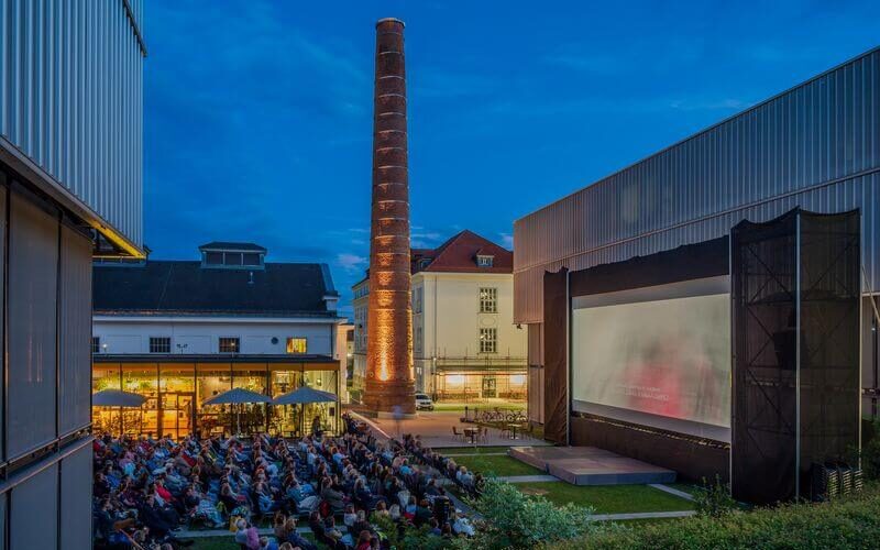 Eine große Gruppe von Menschen sieht sich in der Abenddämmerung einen Film auf einer Freiluftleinwand in einem Innenhof an, der von modernen Gebäuden und einem hohen Backsteinschornstein umgeben ist.