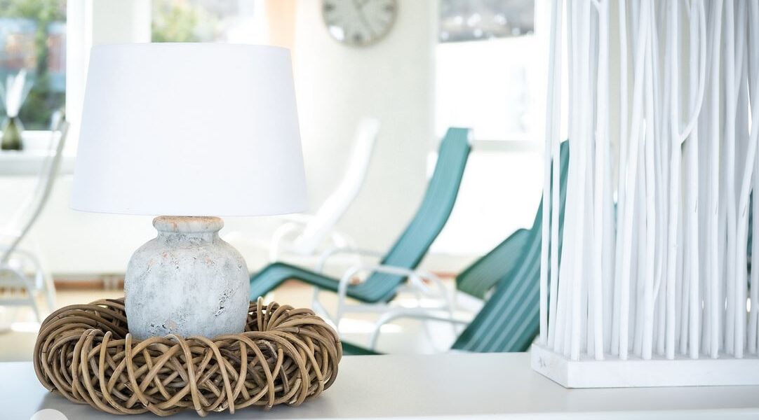 Eine Keramiktischlampe mit weißem Schirm sitzt in einem geflochtenen Rattansockel auf einer weißen Fläche; im hellen Hintergrund sind Liegestühle und eine Uhr zu sehen.