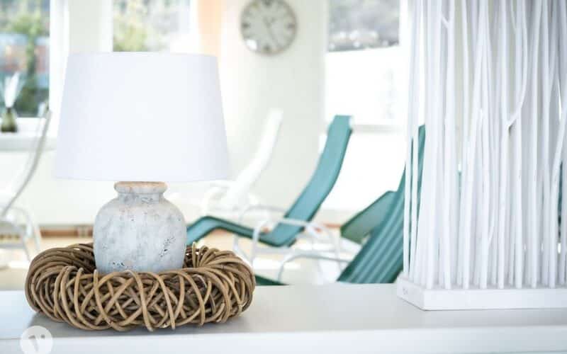 Eine Keramiktischlampe mit weißem Schirm sitzt in einem geflochtenen Rattansockel auf einer weißen Fläche; im hellen Hintergrund sind Liegestühle und eine Uhr zu sehen.