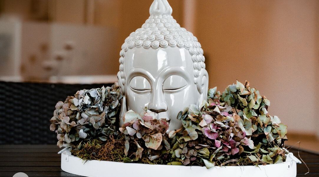 Ein weißer Keramik-Buddha-Kopf, umgeben von getrockneten Hortensienblüten und Moos, ausgestellt auf einem runden Tablett auf einer dunklen Holzoberfläche.