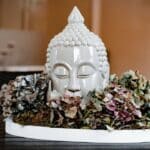 Ein weißer Keramik-Buddha-Kopf, umgeben von getrockneten Hortensienblüten und Moos, ausgestellt auf einem runden Tablett auf einer dunklen Holzoberfläche.