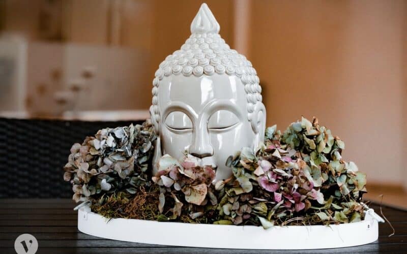 Ein weißer Keramik-Buddha-Kopf, umgeben von getrockneten Hortensienblüten und Moos, ausgestellt auf einem runden Tablett auf einer dunklen Holzoberfläche.