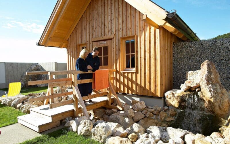 Zwei Personen stehen auf einer Holzrampe vor einer kleinen Holzhütte und blicken auf einen Steingarten mit einem Wasserfall.