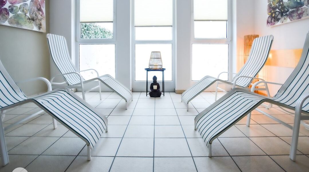 Vier weiße Loungesessel mit blauen Streifen stehen in einem hellen, gefliesten Raum mit großen Fenstern und dekorativer Wandkunst. In der Mitte steht ein kleiner Tisch mit einer Lampe.