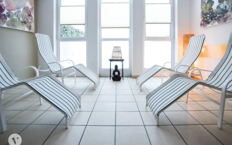 Vier weiße Loungesessel mit blauen Streifen stehen in einem hellen, gefliesten Raum mit großen Fenstern und dekorativer Wandkunst. In der Mitte steht ein kleiner Tisch mit einer Lampe.