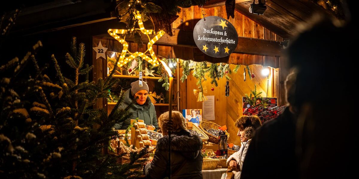 Die Menschen stehen an einem warm beleuchteten, mit Sternen und Grünzeug geschmückten Holzmarktstand und kaufen abends bei einem Verkäufer Backwaren.