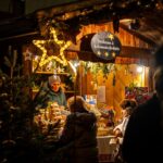 Die Menschen stehen an einem warm beleuchteten, mit Sternen und Grünzeug geschmückten Holzmarktstand und kaufen abends bei einem Verkäufer Backwaren.